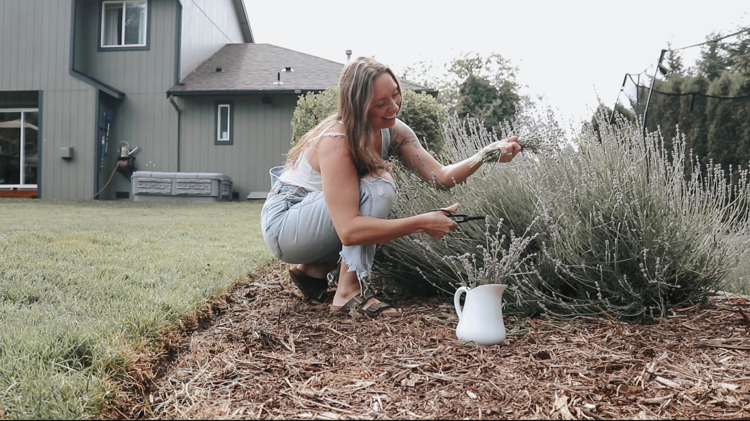 Christian homemaker wearing white tank top and denim overalls crouches in a garden, trimming lavender plants with scissors, with a white watering can placed beside her.